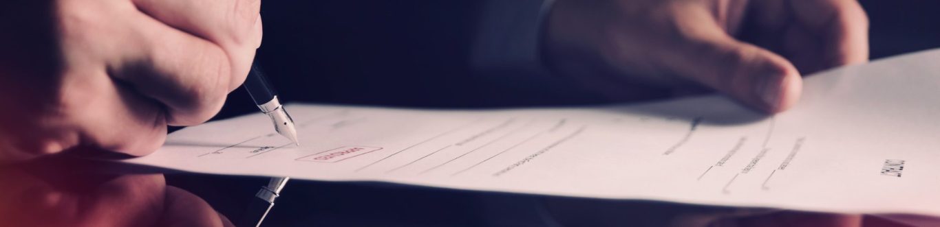Man’s hand signing a document on a desk with a red surface.